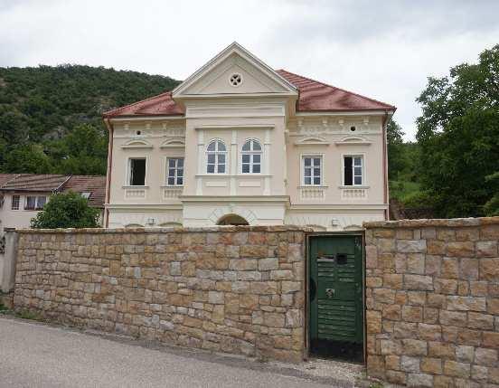 A szerény, nyeregtetős-oromfalas házak között emelt épület az utcai fronton emeletesen, hátul pedig fölszintesen képzett tömeget.