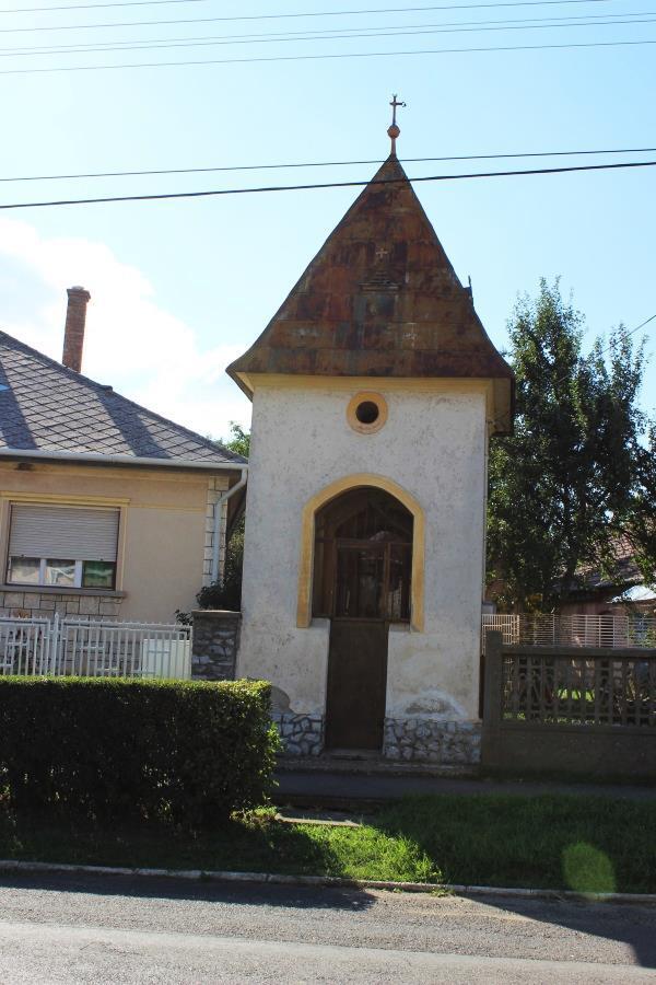 Utcafront vonalában, lakóház portájának szélén álló, kisméretű, gúlatetős kőkápolnácska, utcára