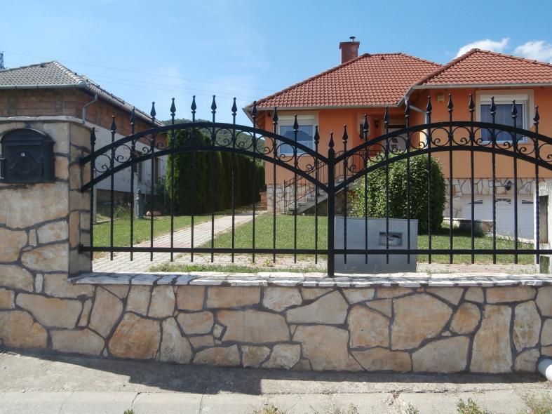 Áttört kovácsoltvas kerítés terméskő burkolatú alapzaton.